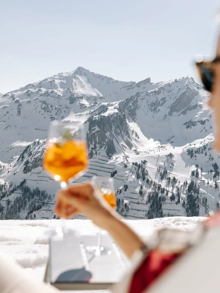 Twee glazen Aperol Spritz die geproost worden, met een besneeuwd bergmassief op de achtergrond.