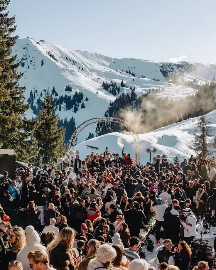 Een volle après-ski aan de voet van een besneeuwde piste, met een podium en de bergen erachter.