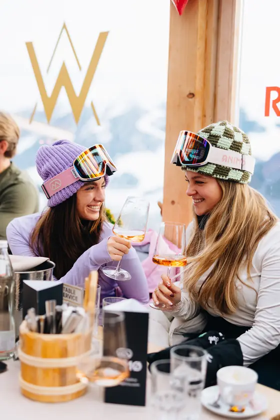 Twee vrouwen met skibril op hun muts proosten met een drankje aan een houten tafel.