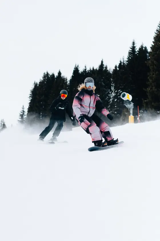 Een snowboarder en een skiër glijden over een besneeuwde piste langs dennenbomen.