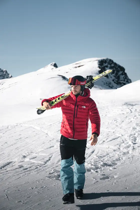 Een skiër in een rode jas loopt met de ski's over zijn schouder over een bergtop.