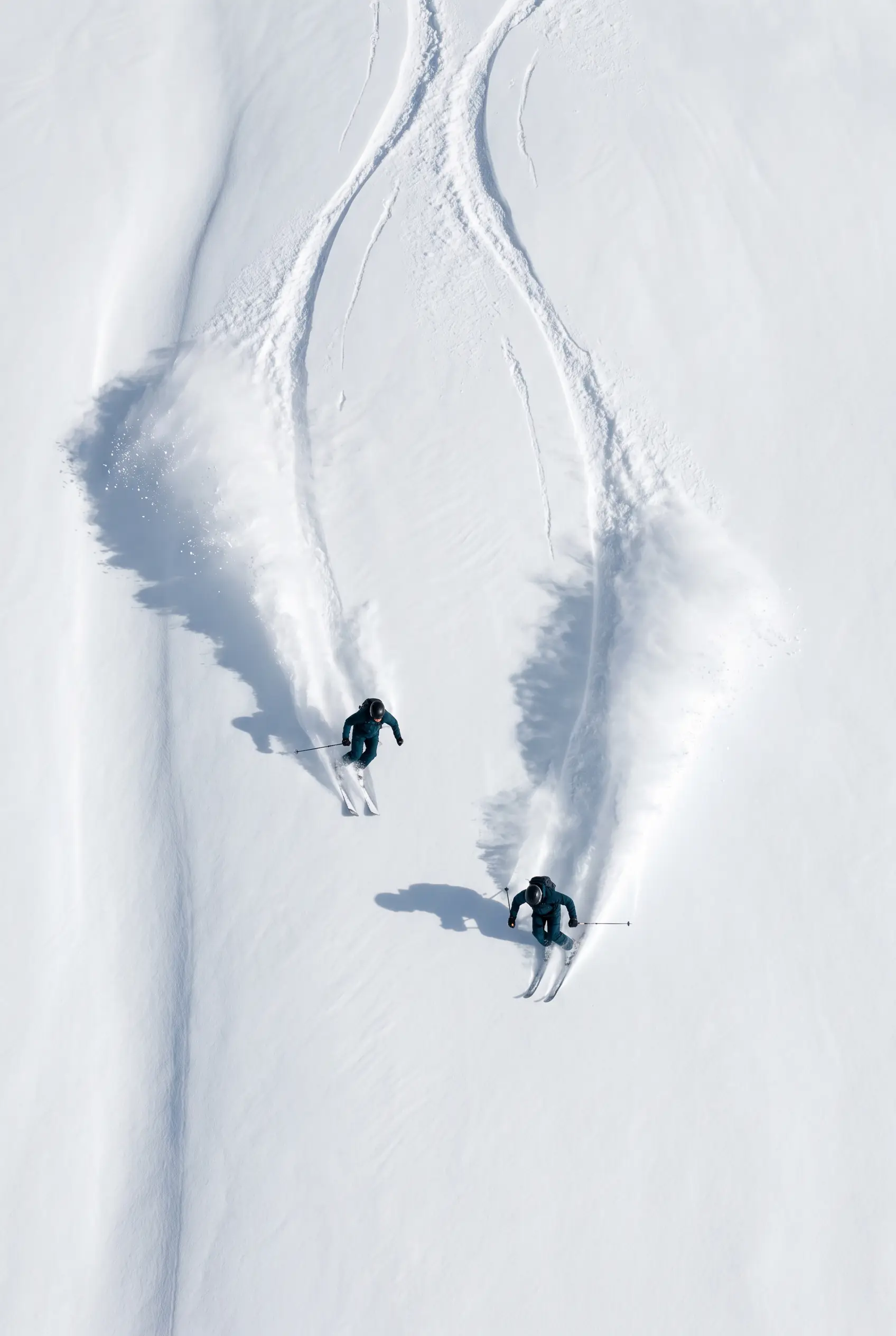 Twee skiërs naast elkaar door verse poedersneeuw, van bovenaf gezien, met twee pluimen sneeuw achter zich.