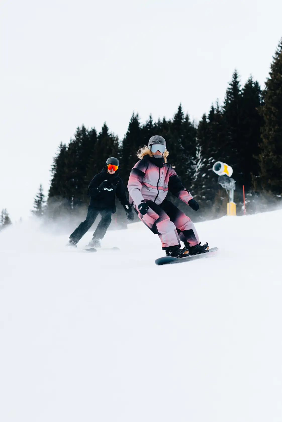 Een snowboarder en een skiër met een camera glijden over een besneeuwde piste langs dennenbomen.
